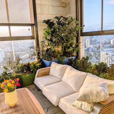 עיצוב גינות יוקרה במרפסת עיצוב גינה במרפסת שמש עם פינת ישיבה לבנה, שולחן עץ ואדניות מעוצבות בצבע ירוק וכחול.