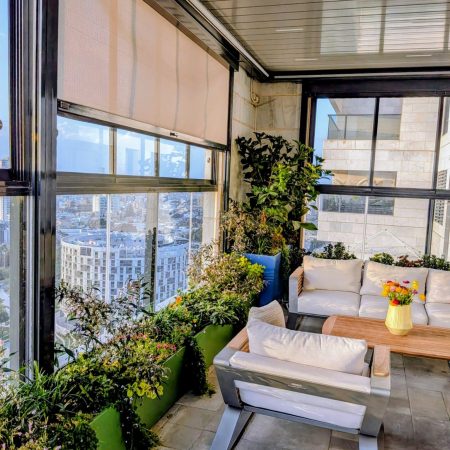 מרפסת סגורה גינת חורף עיצוב גינה במרפסת סגורה עם חלונות ויטרינה גדולים, וילונות גלילה ואדניות ירוקות לאורך המעקה.